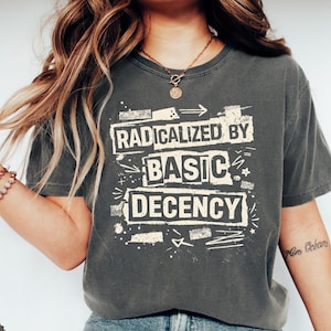 May include: Dark grey t-shirt featuring the text "RADICALIZED BY BASIC DECENCY" in a distressed white font. The shirt has a vintage, worn appearance. The model is wearing a gold necklace and bracelets.