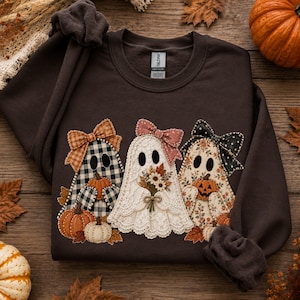 May include: A dark chocolate brown crewneck sweatshirt featuring a graphic of three ghosts. The left ghost has a black and white buffalo check pattern with an orange bow. The center ghost is white lace with a pink bow and flowers. The right ghost has a floral pattern with a black polka dot bow.