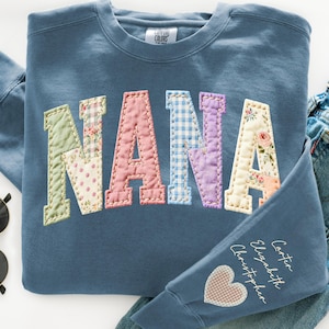 Könnte beinhalten: Ein blaues Sweatshirt mit dem Wort "NANA" in Patchwork-Stoffbuchstaben appliziert. Die Buchstaben sind in Pastellfarben und -mustern gehalten. Der Ärmel hat eingestickte Namen und einen Herzflicken.
