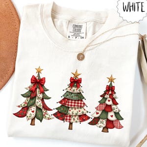 Peut inclure: Sweat-shirt blanc Comfort Colors avec trois sapins de Noël ornés de nœuds rouges, d'étoiles dorées et de motifs patchwork rouges, verts et blancs. Un collier doré est drapé sur le vêtement. Le mot "WHITE" est dans un cercle blanc.