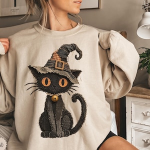 Könnte beinhalten: Ein sandfarbenes Sweatshirt mit Rundhalsausschnitt und einer Grafik einer schwarzen Katze, die einen abgenutzten Hexenhut mit kariertem Flicken und ein goldenes Glockenhalsband trägt. Die Katze hat große orangefarbene Augen und eine Textur.