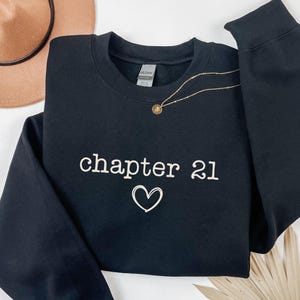 Peut inclure: Sweat-shirt noir avec l'inscription "chapter 21" et un cœur blanc. Un collier doré avec un pendentif est posé sur le sweat-shirt. Un chapeau marron et une feuille de palmier séchée sont en arrière-plan.