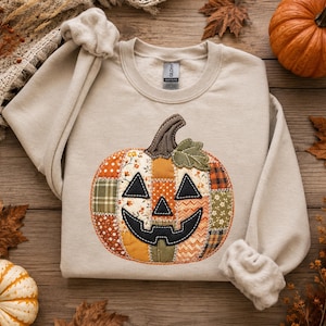 Peut inclure: Un sweat-shirt à col rond couleur sable orné d'une grande applique de citrouille centrale. La citrouille est composée de divers empiècements en tissu orange, vert et marron à motifs floraux et écossais, avec un visage de citrouille noir.