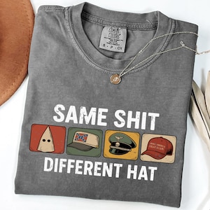 Könnte beinhalten: Graues T-Shirt mit dem Text "SAME SHIT DIFFERENT HAT" und Illustrationen einer KKK-Haube, eines Konföderiertenhuts, eines Militärhuts und einer "Make America Great Again"-Kappe. Eine silberne Halskette mit Anhänger ist ebenfalls zu sehen.