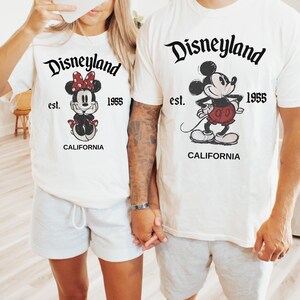 May include: White t-shirts with Disneyland graphics. One shirt features Minnie Mouse with a red bow, and the other features Mickey Mouse in red shorts. Both shirts have "Disneyland est. 1955 CALIFORNIA" printed in black.