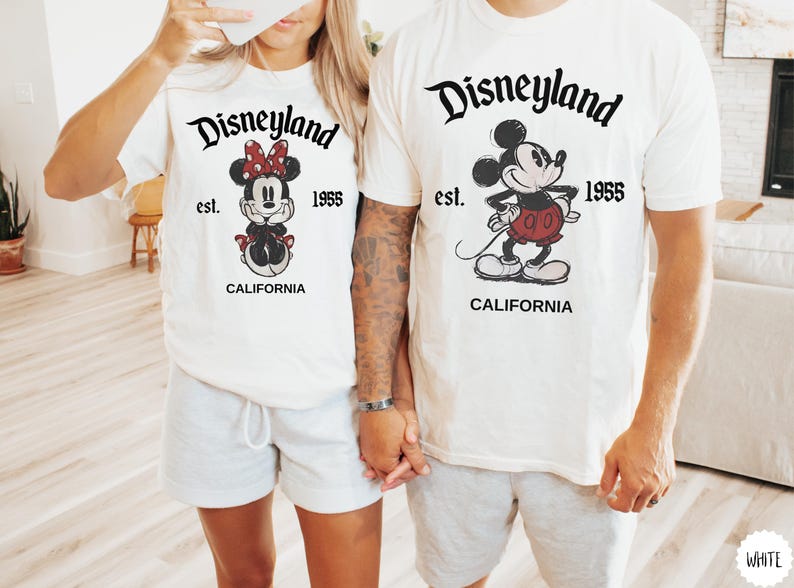May include: White t-shirts with Disneyland graphics. One shirt features Minnie Mouse with a red bow, and the other features Mickey Mouse in red shorts. Both shirts have "Disneyland est. 1955 CALIFORNIA" printed in black.