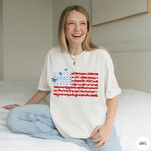 May include: White t-shirt featuring a floral American flag design. The flag's stars are represented by white flowers on a blue background, and the stripes are red and white flowers. Three blue butterflies are scattered across the design.
