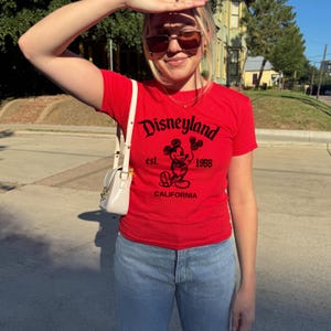 May include: A red t-shirt with the word "Disneyland" in black script, featuring a Mickey Mouse graphic and the text "est. 1955 CALIFORNIA". The shirt is paired with light blue jeans and a white purse.