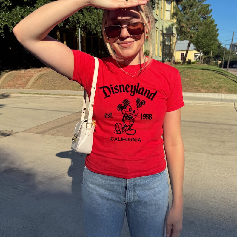 May include: A red t-shirt with the word "Disneyland" in black script, featuring a Mickey Mouse graphic and the text "est. 1955 CALIFORNIA". The shirt is paired with light blue jeans and a white purse.