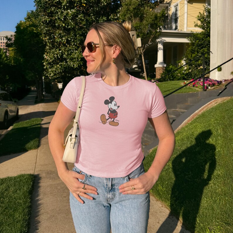 Peut inclure: Un t-shirt rose clair orn&eacute; d'un motif Mickey Mouse classique. Le t-shirt &agrave; manches courtes est associ&eacute; &agrave; un jean bleu clair. La personne porte des lunettes de soleil et un sac &agrave; main blanc. L'arri&egrave;re-plan comprend des arbres et un b&acirc;timent.