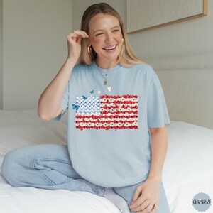 May include: Light blue t-shirt featuring a floral American flag design. The flag is composed of red and white flowers with a blue field of white flowers. Butterflies are scattered around the flag. The shirt is a relaxed fit.