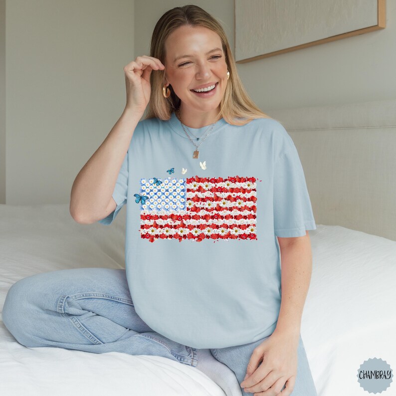 May include: Light blue t-shirt featuring a floral American flag design. The flag is composed of red and white flowers with a blue field of white flowers. Butterflies are scattered around the flag. The shirt is a relaxed fit.