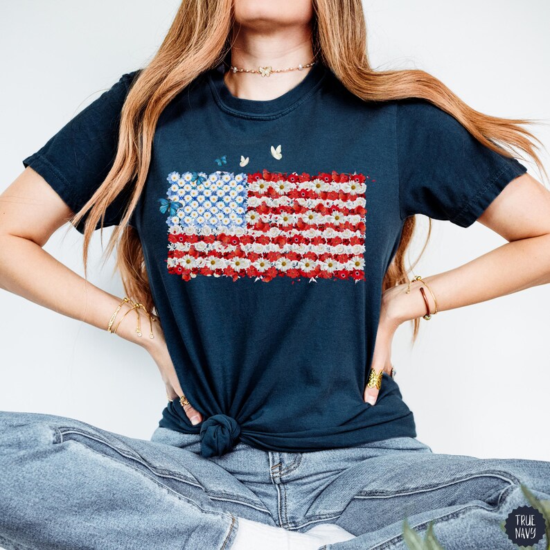 May include: Navy blue t-shirt featuring a floral American flag design. The flag is composed of white and red flowers with a blue square of white flowers and butterflies in the upper left corner. The shirt is tied at the waist.