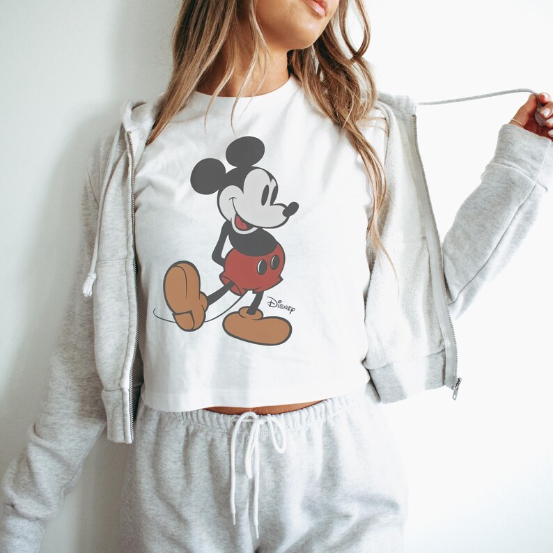 May include: White crop top featuring a classic Mickey Mouse graphic. The cartoon character is depicted in black, red, and brown, with the Disney logo below. The top is part of a casual outfit.