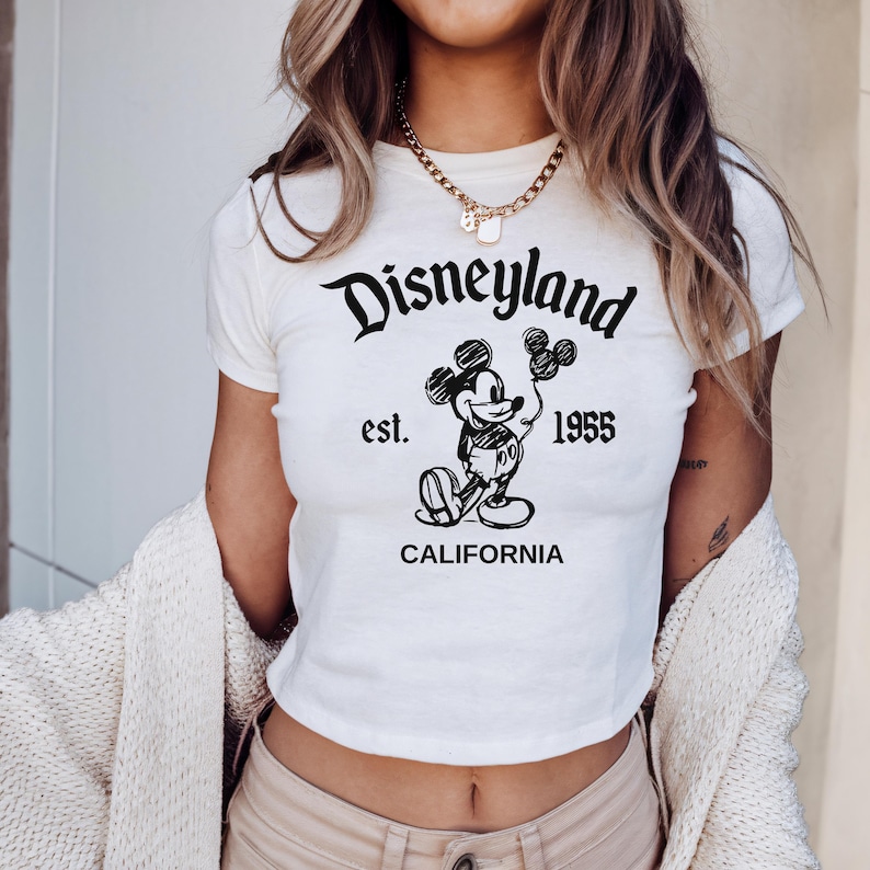 May include: White crop top featuring a black graphic of Mickey Mouse holding balloons, with the text "Disneyland est. 1955 CALIFORNIA". The shirt has short sleeves and a round neckline.