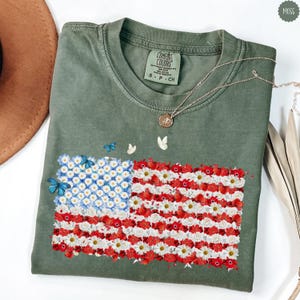 May include: A moss green t-shirt featuring a floral American flag design. The flag is composed of white daisies, red flowers, and blue flowers. The shirt has a Comfort Colors tag and is adorned with a silver necklace. Small butterflies are scattered around the flag.