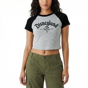 May include: A gray and black raglan crop top with the word "Disneyland" in black script. The shirt features a Mickey Mouse graphic and the text "EST. 1955". The model is wearing olive green cargo pants.