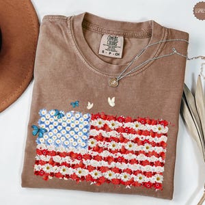 May include: A brown Comfort Colors t-shirt featuring a floral American flag design. The flag is made of white and red flowers with blue flowers in the corner. Butterflies are scattered around the flag. A silver necklace is draped across the shirt.