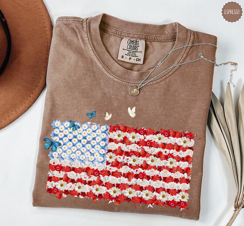May include: A brown Comfort Colors t-shirt featuring a floral American flag design. The flag is made of white and red flowers with blue flowers in the corner. Butterflies are scattered around the flag. A silver necklace is draped across the shirt.