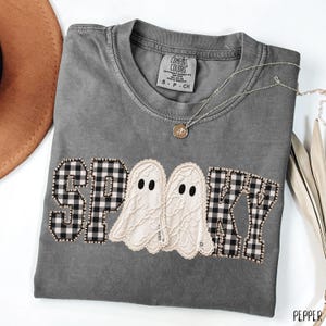 Könnte beinhalten: Ein anthrazitfarbenes Kurzarm-T-Shirt mit dem Wort SPOOKY als Applikation auf der Brust. Die Buchstaben S, P, K und Y bestehen aus schwarz-weiß kariertem Stoff, während die beiden O's als weiße Geister aus Spitze gestaltet sind.