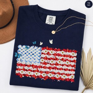 May include: A navy blue t-shirt featuring a floral American flag design. The flag is composed of white daisies, red flowers, and blue butterflies. A gold necklace and a brown hat are visible in the background. The shirt has a Comfort Colors label.