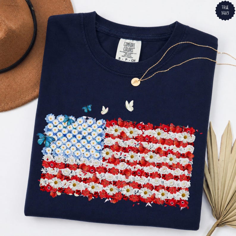 May include: A navy blue t-shirt featuring a floral American flag design. The flag is composed of white daisies, red flowers, and blue butterflies. A gold necklace and a brown hat are visible in the background. The shirt has a Comfort Colors label.