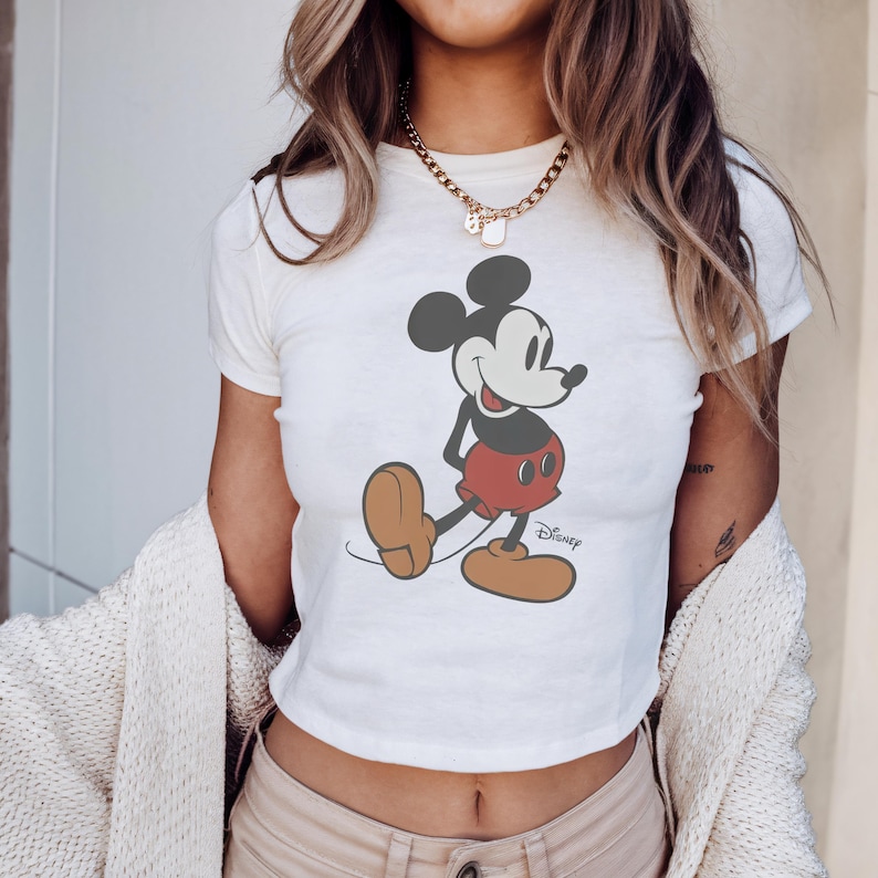 May include: White crop top featuring a classic Mickey Mouse graphic. The cartoon character is depicted in his iconic pose, wearing red shorts, brown shoes, and black gloves. The Disney logo is printed below the character.