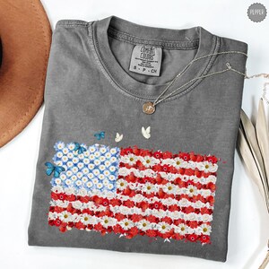 May include: A gray Comfort Colors t-shirt featuring a floral American flag design. The flag is composed of white daisies, red flowers, and blue butterflies. A silver necklace with a pendant and small butterflies are also visible.