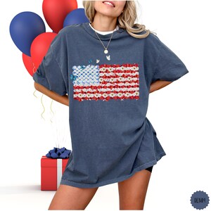 May include: A denim blue t-shirt featuring a floral American flag design. The flag's stripes are made of red and white flowers, with a blue flower field in the corner. The shirt is part of a patriotic collection.