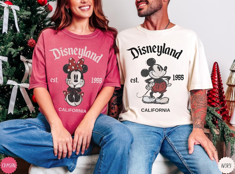 May include: Two t-shirts with Disneyland graphics. One is crimson with a Minnie Mouse design, the other is ivory with Mickey Mouse. Both shirts have "Disneyland est. 1955 California" printed on them.