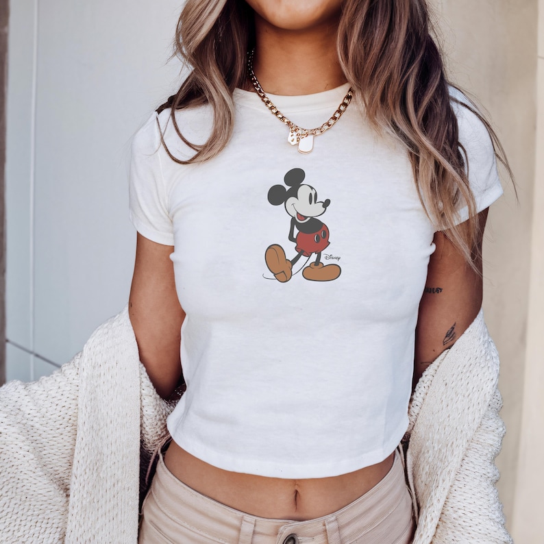 Peut inclure: Haut court blanc avec un motif de Mickey Mouse. Le personnage de dessin anim&eacute; est repr&eacute;sent&eacute; dans sa pose embl&eacute;matique, portant un short rouge, des chaussures marron et des gants noirs. Le logo Disney est imprim&eacute; en dessous.