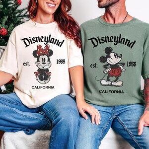 May include: Two t-shirts, one ivory and one moss green, featuring Disneyland graphics. The ivory shirt displays Minnie Mouse, and the green shirt features Mickey Mouse. Both shirts have the text "Disneyland est. 1955 CALIFORNIA".