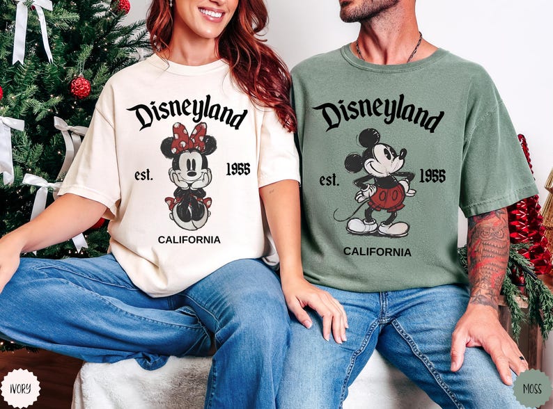 May include: Two t-shirts, one ivory and one moss green, featuring Disneyland graphics. The ivory shirt displays Minnie Mouse, and the green shirt features Mickey Mouse. Both shirts have the text "Disneyland est. 1955 CALIFORNIA".
