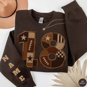 May include: A dark brown crewneck sweatshirt featuring a large number 18 on the front, constructed from various brown and tan fabric patches with frayed edges. The number 18 includes star and football lace motifs. The word NAME is printed on the sleeve.