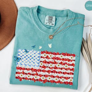 May include: A seafoam green t-shirt featuring an American flag design made of white and red flowers, with blue butterflies. The shirt has a Comfort Colors tag and a gold necklace. The flag design is made of white daisies and red flowers.