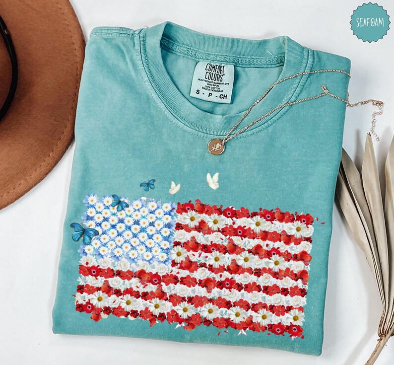 May include: A seafoam green t-shirt featuring an American flag design made of white and red flowers, with blue butterflies. The shirt has a Comfort Colors tag and a gold necklace. The flag design is made of white daisies and red flowers.