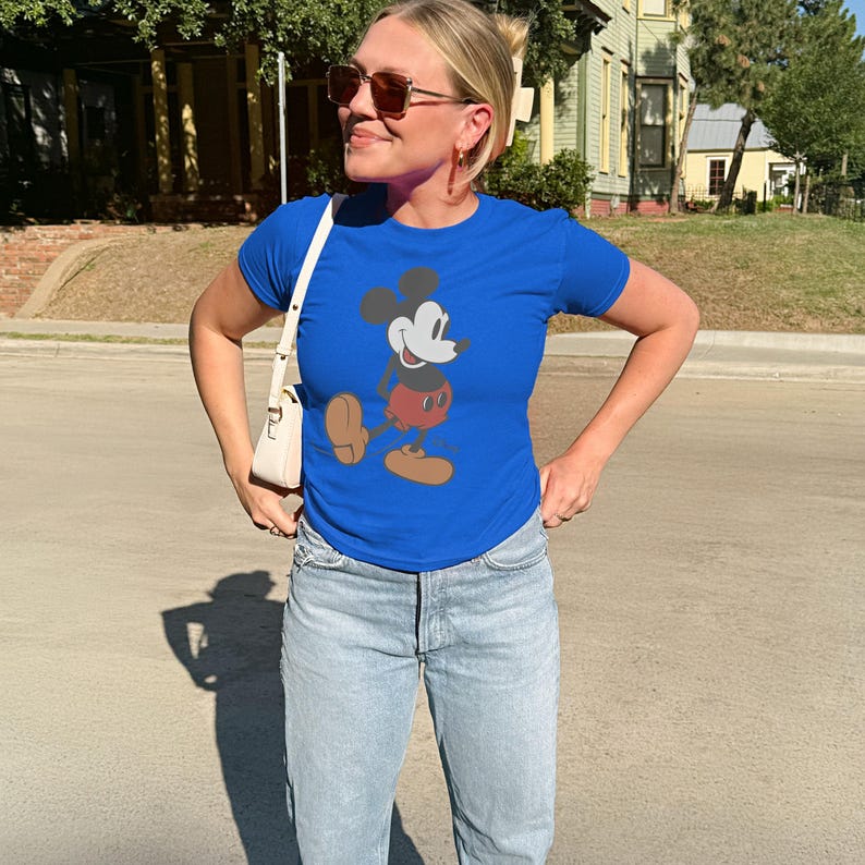 May include: A royal blue t-shirt featuring a graphic of Mickey Mouse. The cartoon character is depicted in his classic pose, with a white face, black ears, red shorts, and brown shoes. The shirt is paired with light-wash denim jeans.