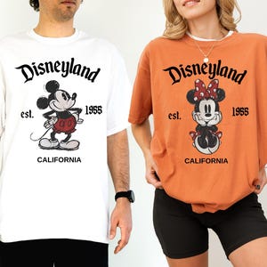 May include: Two t-shirts, one white and one orange, featuring the Disneyland logo and Mickey and Minnie Mouse. The white shirt has Mickey Mouse standing, while the orange shirt has Minnie Mouse sitting. Both shirts have "est. 1955" and "CALIFORNIA" printed below the characters.