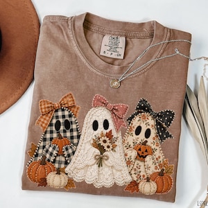 May include: A brown short-sleeve t-shirt featuring a graphic of three ghosts. The left ghost has a black and white buffalo check pattern with an orange bow and pumpkins. The middle ghost is white lace with a pink bow and flowers. The right ghost has a floral pattern with a black polka dot bow and pumpkins.