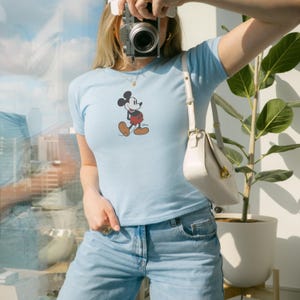 Peut inclure: T-shirt bleu clair avec un motif Mickey Mouse. Le t-shirt est associ&eacute; &agrave; un jean en denim bleu clair. Un sac &agrave; main blanc avec un fermoir dor&eacute; est port&eacute; &agrave; l'&eacute;paule. Un appareil photo est tenu devant le visage.