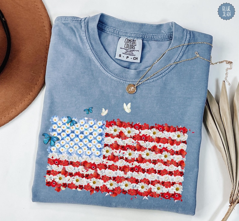 May include: A light blue Comfort Colors t-shirt featuring a floral American flag design. The flag is made of white daisies and red flowers, with blue butterflies. A gold necklace with a pendant rests on the shirt.