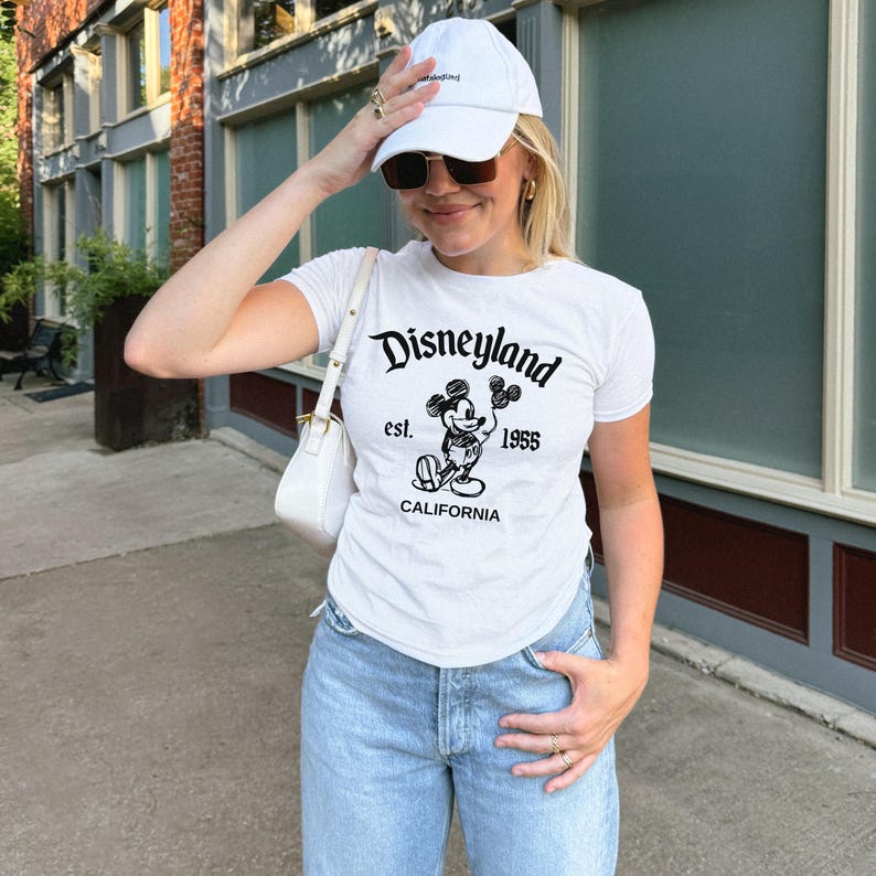 May include: White t-shirt with a black Disneyland graphic featuring Mickey Mouse, est. 1955, and the word California. The person is wearing a white baseball cap and sunglasses.