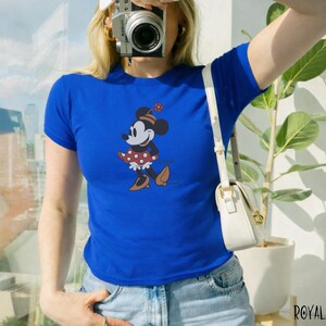 Puede incluir: Camiseta azul real con un gr&aacute;fico de Minnie Mouse. El personaje de dibujos animados lleva una falda roja de lunares, zapatos marrones y un sombrero con una flor. La camiseta tiene mangas cortas y cuello redondo.