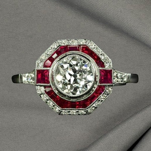 May include: A platinum ring with a large, round, brilliant-cut diamond center stone. The ring features a halo of smaller diamonds and red rubies, set in an octagonal shape. The band is also adorned with diamonds.