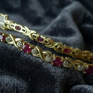 Könnte beinhalten: Nahaufnahme von zwei goldenen Armbändern mit abwechselnd ovalen Rubin-Edelsteinen und kleinen Diamanten. Die Armbänder haben ein kompliziertes, verschlungenes Design und sind vor einem dunklen, strukturierten Hintergrund platziert.