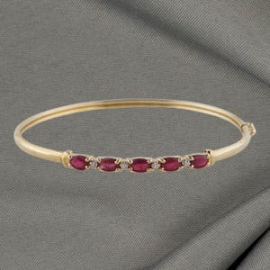 May include: A gold bangle bracelet featuring six oval, deep red gemstones and small, clear accent stones. The bracelet is set against a grey background.