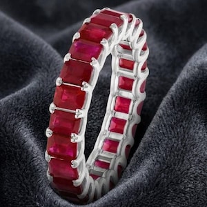 May include: A silver ring featuring a continuous row of rectangular, deep red gemstones. The gemstones are set closely together, creating a full band of color. The ring is displayed on a dark, textured surface.