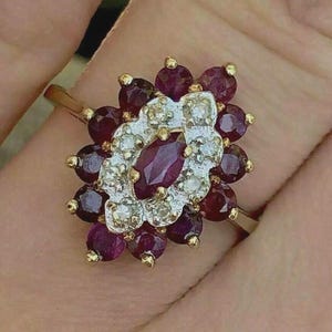 May include: A gold ring featuring a cluster of gemstones. The ring's centerpiece is an oval-cut ruby, surrounded by smaller, round rubies and diamonds. The gemstones are set in a floral pattern, with gold accents.