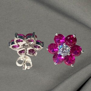 May include: A pair of flower-shaped stud earrings. Each earring features six magenta-colored gemstones arranged around a central, clear gemstone. The earrings are set in a silver-colored metal, with a post and butterfly backing.