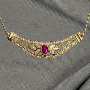 May include: A gold-toned necklace with a central oval ruby and surrounding clear gemstones. The necklace features a delicate filigree design and is suspended from a fine chain. The overall design is symmetrical and elegant.